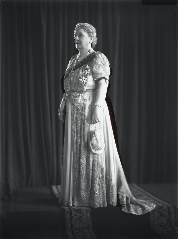 Schwarzes und weißes Foto von Queen Elizabeth II auf einer Bühne mit einem Vorhanghintergrund, die eine königliche Robe mit detaillierten Verzierungen und einem langen Rock trägt, mit hochgesteckten Haaren und einem sanften Gesichtsausdruck zur Seite schaut.