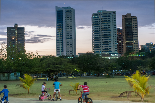 Kinder auf Fahrrädern in einem Park bei Sonnenuntergang mit saftigem Grün und Bäumen, gegen einen Himmel aus Orangen, Pinks und Purpurs, und skylined Hochhäuser im Hintergrund.