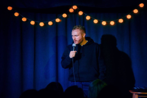 Ein Mann steht auf einer hell erleuchteten Bühne und hält ein Mikrofon während einer Stand-up-Comedy-Show in der Hand, mit einem Tisch voller Gegenstände rechts und einem sichtbaren Publikum darunter.