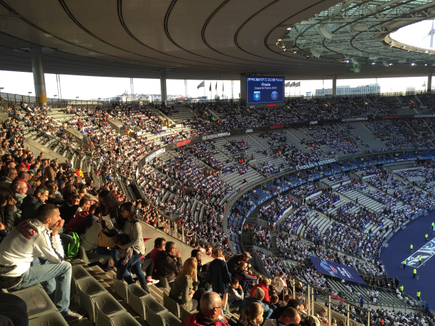 Große Zuschauermenge in einem Stadion bei einem Fußballspiel, mit einer Bühne rechts, Fahnen, Stangen, einem Bildschirm und der Allianz Arena in München, Deutschland im Hintergrund.