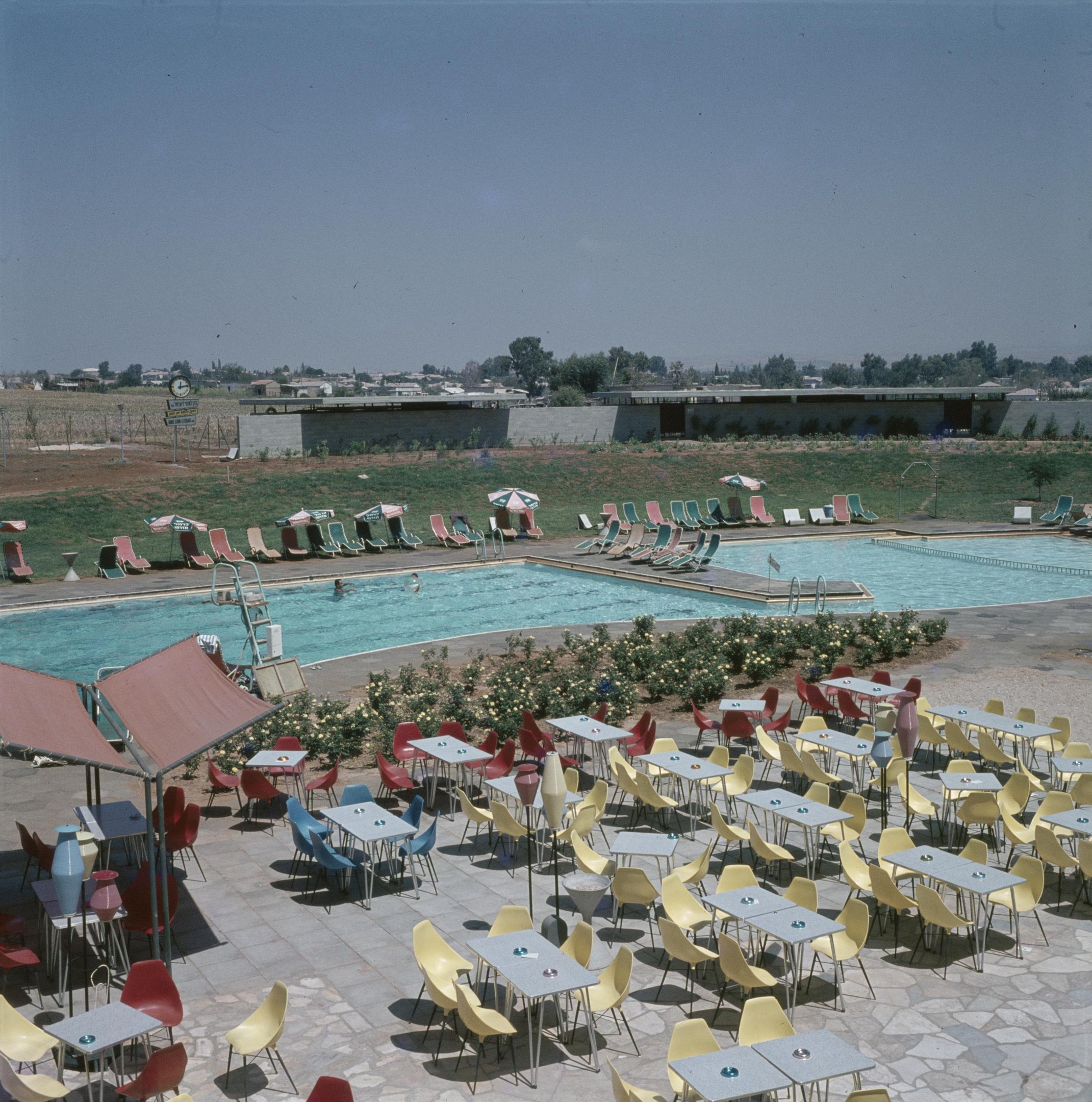 Ein Freibad umgeben von Tischen, Stühlen, Blumenpflanzen, Sonnenschirmen, Pfählen, einem Zaun, einer Wand, einer Baumgruppe, einigen Gebäuden unter einem bewölkten Himmel.