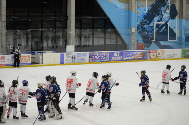 Junge Menschen in Helmen und Hockey-Ausrüstung spielen Eis-hockey auf einer Indoor-Eisfläche, mit einem Zuschauer links und Bannern, Säulen und Glaswänden im Hintergrund.