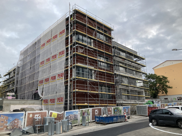 Großes Gebäude in Bau mit Gerüst, umgeben von Bäumen, Straßeninfrastruktur und Fahrzeugen, vor einem bewölkten Himmel.