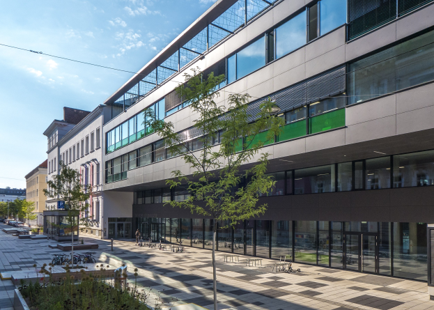 Großes modernes Gebäude mit vielen Fenstern, umgeben von Bäumen und Grünflächen, mit Menschen und Fahrzeugen in der Nähe, unter einem bewölkten Himmel, als das neue deutsche Institut für Technologie-Hauptquartier in Berlin identifiziert.
