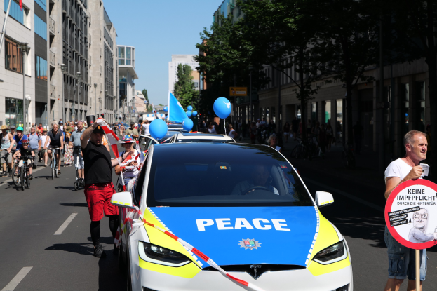 Eine Gruppe von Menschen, die auf Fahrrädern eine Straße entlangfahren, mit einem Polizeiwagen im Vordergrund, einer Person, die eine Tafel mit einem Bild und Text hält, und Gebäuden, Bäumen und einem klaren Himmel im Hintergrund.