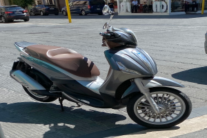 Ein Motorroller am Straßenrand mit einem Helm darauf, mit Gebäuden, Bäumen, Pfählen, Menschen, Stühlen, Tischen und anderen Gegenständen im Hintergrund.
