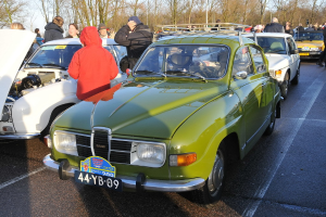 Mehrere Autos sind mit einer Gruppe von Menschen in der Nähe geparkt, die meisten tragen Jacken, vor einem Hintergrund von Bäumen, Pfählen und dem Himmel.
