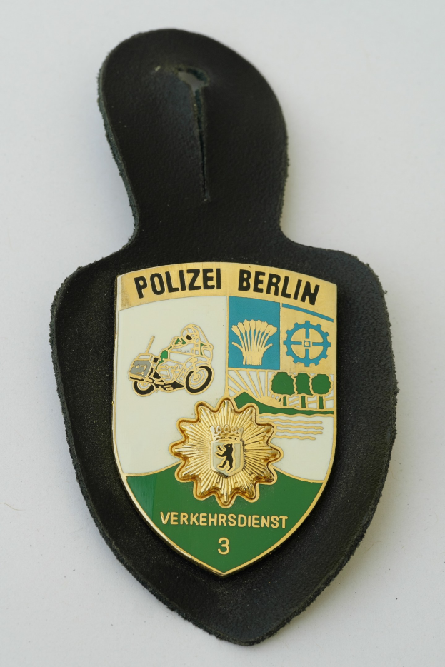 Ein schildförmiges Polizeibadgeb mit der Aufschrift "Polizei Berlin" auf einem blauen Hintergrund, einem weißen Rand, einem weißen zentralen Stern und schwarzer fetter Schrift, das auf einer Oberfläche liegt.