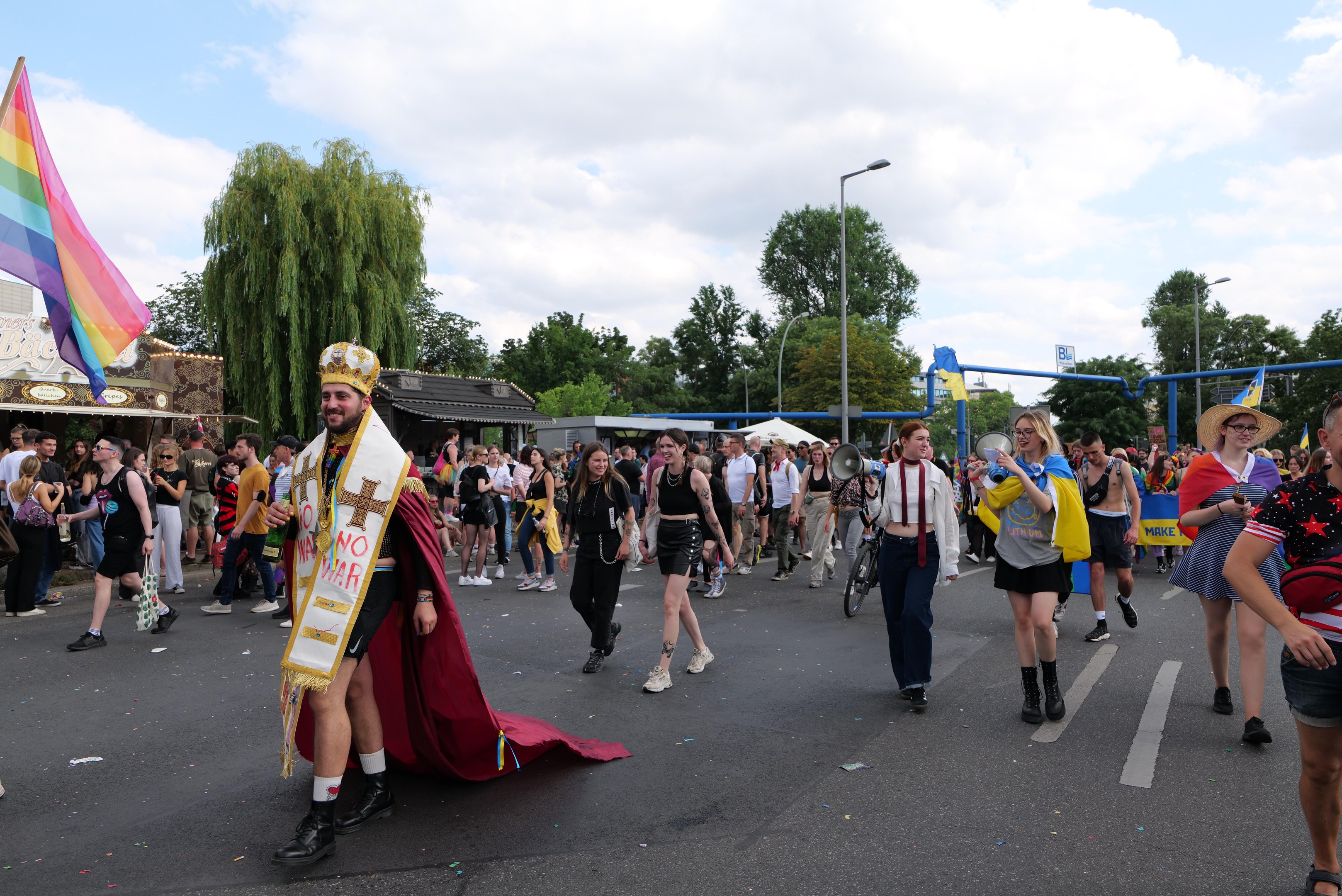 Eine Gruppe von Menschen marschiert bei der Gay Pride Parade 2018 mit einer Regenbogenflagge und Musikinstrumenten, während im Hintergrund Laternenpfähle, Bäume, Schuppen und ein bewölkter Himmel zu sehen sind.