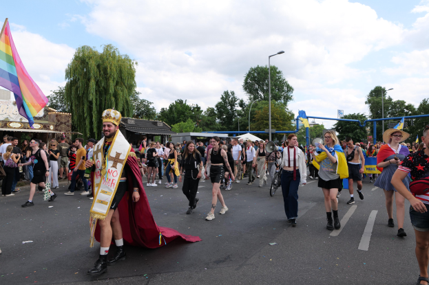 Eine Gruppe von Menschen marschiert bei der Gay Pride Parade 2018 mit einer Regenbogenflagge und Musikinstrumenten, während im Hintergrund Laternenpfähle, Bäume, Schuppen und ein bewölkter Himmel zu sehen sind.