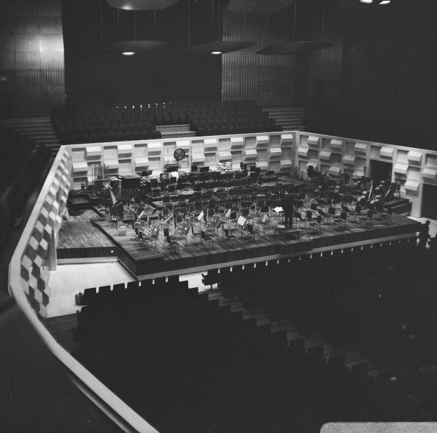 Ein Schwarz-Weiß-Foto eines großen Auditoriums mit einer Bühne in der Mitte, umgeben von Stühlen und beleuchtet von Deckenlampen, auf der ein paar Menschen Musikinstrumente spielen.
