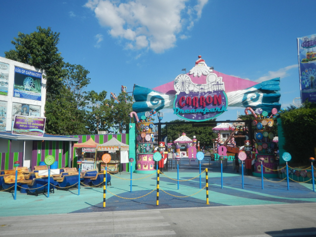 Kirmesfahrgeschäft am Parkeingang mit Straße darunter, eingezäuntem Bereich, Buden, Bannern, Bäumen und bewölktem Himmel im Hintergrund.