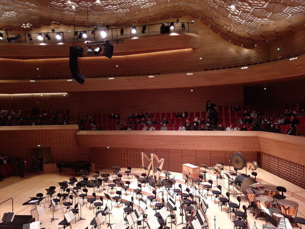 Ein großer Auditorium mit Musikinstrumenten, Stühlen, Ständern und Menschen, mit beleuchteter Decke, darstellend den Hauptsaal der Berliner Philharmonie.