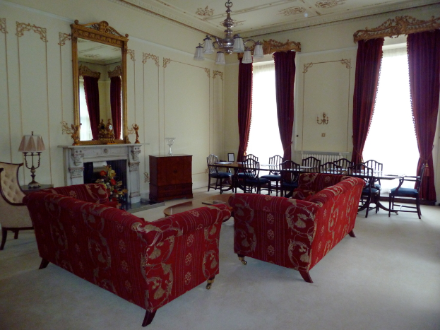 Zwei rote Sofas in einem Wohnzimmer, ein kleiner Teetisch mit einem Buch darauf, ein wandhängender Spiegel, eine Lampe auf einem Tisch, ein Blumenstrauß vor einem Kamin, ein Esstisch mit Stühlen, von der Decke hängende Lampen, Vorhänge an einer Stange und ein leerer Stuhl.