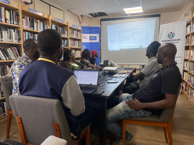 Eine Gruppe von Menschen sitzt um einen Tisch mit Laptops und Kabeln, die an einem Workshop über Technologie in der Bildung teilnehmen, mit einem Projektor, Bannern, Bücherregalen und einer Klimaanlage im Raum.