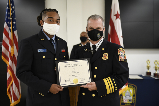 Zwei uniformierte Feuerwehrleute, darunter der District of Columbia Fire Chief und der Assistant Fire Chief, stehen nebeneinander und halten ein Zertifikat vor einem mit blauen Schilden und anderen Gegenständen bedeckten Tisch, mit Flaggen und einer Wand im Hintergrund.