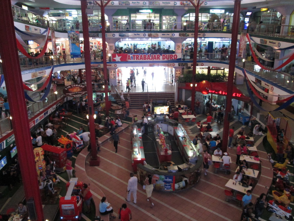 Eine belebte Einkaufsstraßenszene mit Säulen, Menschen, Tischen, Fliesenboden, Flaggen, Geschäften, einem Banner, Treppen und einem Namensschild.