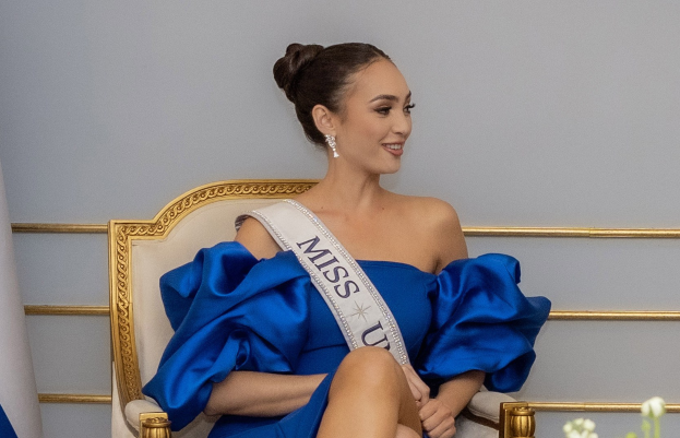 Frau in einem blauen Kleid mit einem "Miss Universe 2019" Band, die auf einem Stuhl sitzt und von Blumen umgeben ist.