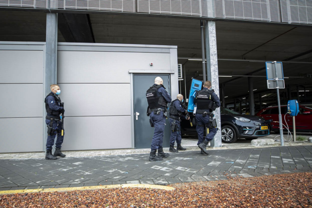 Gruppe von Polizeibeamten, die vor einem Gebäude mit einer Tür in einem Parkplatz stehen, umgeben von Fahrzeugen, Pfosten, Brettern und verstreuten Steinen.