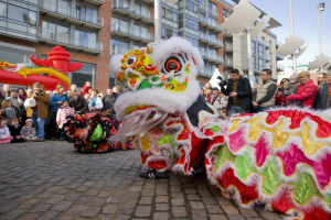 Ein lebendiges chinesisches Neujahrsfest in Amsterdam mit einem Löwen tanzen und einer Menge Schaulustiger, einige machen Fotos, vor einer Kulisse aus Gebäuden, Laternenmasten und einem klaren blauen Himmel.