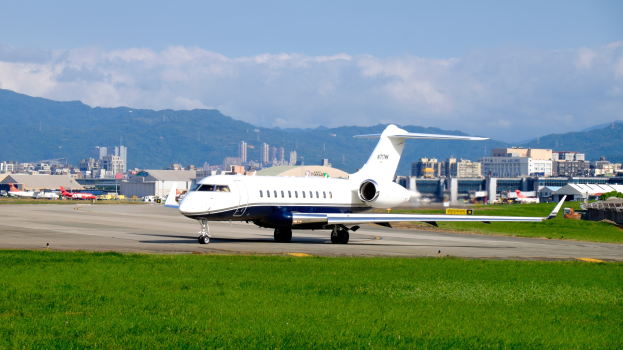 Privater Jet auf einer Flughafenpiste mit anderen Flugzeugen, Gebäuden, Bäumen, Bergen und einem klaren blauen Himmel im Hintergrund.