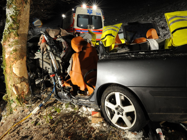 Ein beschädigt aussehendes Auto am Rande einer verschneiten Straße mit einem Krankenwagen im Hintergrund, umgeben von Bäumen.