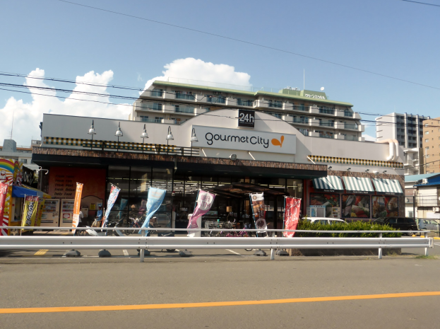 Eine belebte Stadtstraße mit Gebäuden, Straßeneinrichtungsgegenständen, Fahrzeugen, Grünanlagen und einem Restaurant mit Essensauslage im Schaufenster.