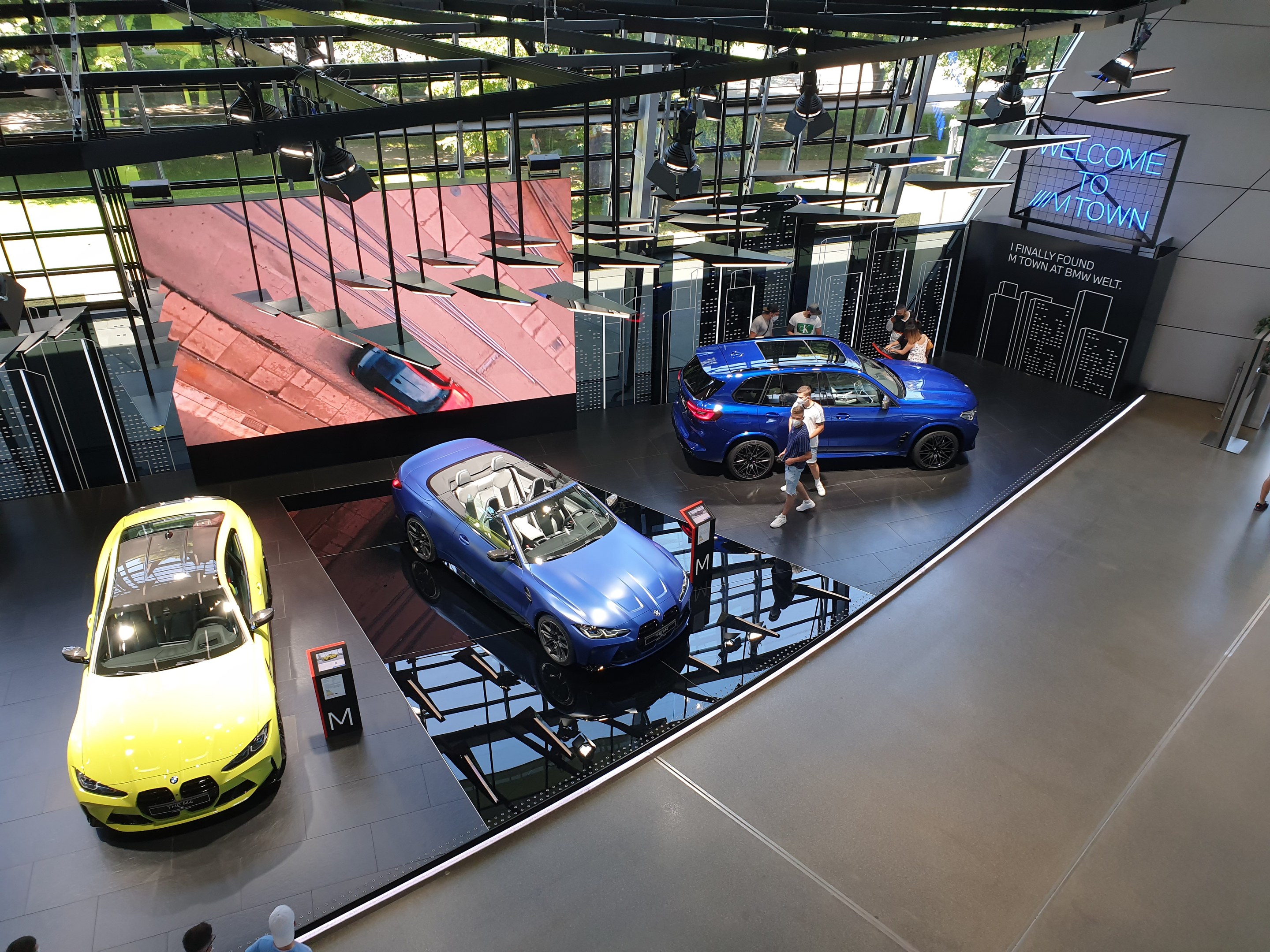 Eine Gruppe von Menschen, die im BMW Museum in München stehen, mit ausgestellt Autos auf der linken Seite und einem Schild auf der rechten Seite, das "BMW Museum München" lautet, mit Blick auf eine Außenansicht von Bäumen, Gebäuden und Himmel durch Glasfenster.