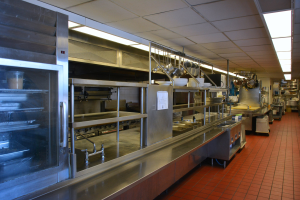 Kommerzielle Küche mit Edelstahl-Theke, elektrischer Deckenbeleuchtung, hängenden Utensilien, einem Waschbecken, Wasserhähnen, Schränken und verschiedenen auf Gestellen angeordneten Gegenständen.