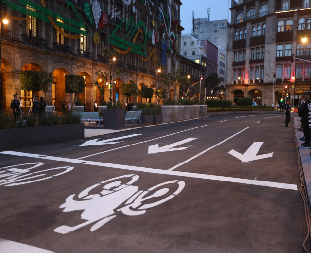 Stadtstraße mit einem ausgewiesenen Fahrradstreifen, umgeben von Gebäuden, Straßenmöbeln, Fußgängern, Grünflächen und einem klaren blauen Himmel.