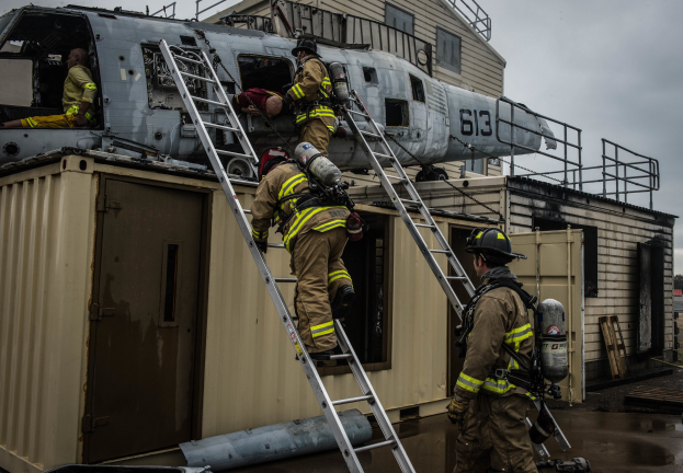 Feuerwehrleute in Uniform und Helmen arbeiten an einem Hubschrauber auf einem Gebäudedach, einer hält einen Sauerstoffzylinder, mit Leitern, einem Container, Bäumen und einem klaren Himmel im Hintergrund.
