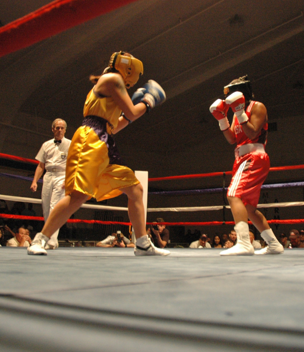 Zwei Boxer im Ring bei einem Kampf mit einem Schiedsrichter und Zuschauern im Hintergrund, einige halten Kameras, unter einer sichtbaren Decke.