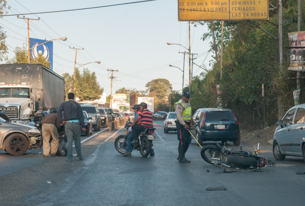 Eine Gruppe von Menschen steht um ein verunglücktes Motorrad auf der Straße herum, mit mehreren Fahrzeugen, darunter ein Lastwagen, und einem Hintergrund aus Bäumen, Masten, Lichtern und Schildern unter dem Himmel.
