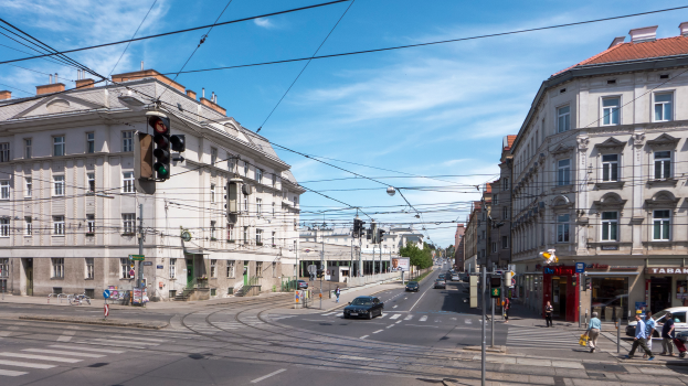 Eine belebte Stadtstraße mit Autos, Fußgängern, Gebäuden, Verkehrsampeln, Schildern, Bäumen und einem bewölkten Himmel.