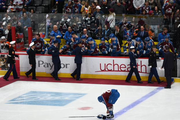 Ein Eishockeyspieler in einer Spielsituation, umgeben von Teamkollegen und Zuschauern, mit Sitzplätzen im Hintergrund und Arena-Banden, die ein Spiel zwischen Colorado Avalanche und San Jose Sharks ankündigen.