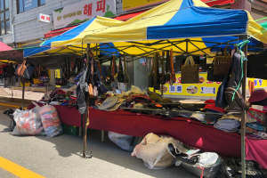 Ein belebter Straßenmarkt mit Zelten, auf denen Taschen, Hüte und andere Artikel ausliegen, mit Gebäuden und Texttafeln im Hintergrund.