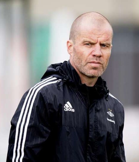 Mann in marineblauer Jacke mit weißen Streifen und weißem Kragen, der auf einem Fußballfeld mit verschränkten Händen und ernster Miene steht.