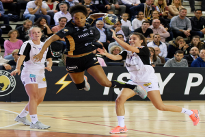 Eine Gruppe von Frauen spielt Handball auf einem Platz, mit einem Ball in der Mitte, während Zuschauer im Hintergrund und ein Banner mit Text und Logos zu sehen sind.