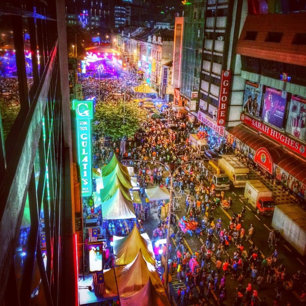 Eine belebte Karnevals-Szene mit einer großen Menge auf der Straße, umgeben von Zelten, Geschäften, Gebäuden und Bannern, mit zahlreichen Lichtern, die den Hintergrundbereich erhellen.