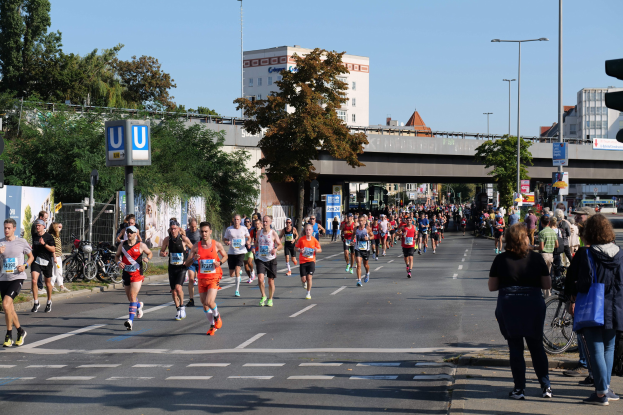 Eine Gruppe von Menschen, die bei einem Marathon auf einer von Bäumen gesäumten Straße mit Laternenmasten, Schildern, Fahrrädern, einem Zaun, Gras, einer Brücke, Gebäuden und einem klaren blauen Himmel laufen.