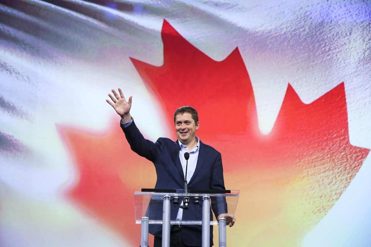 Der kanadische Premierminister Alex Salmond steht vor einem kanadischen Flagge mit Mikrofonen auf einem Podium und lächelt in die Kamera. Im Hintergrund ist ein Bildschirm zu sehen.