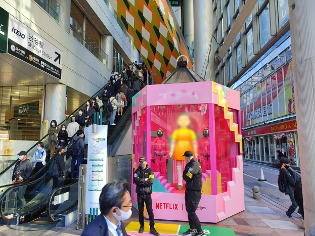 Eine Gruppe von Menschen steht vor einem Gebäude mit Glaswänden, Säulen und Texttafeln, einige tragen Masken, mit einer Rolltreppe im Blick; eine rosafarbene Box in der Mitte zeigt Schaufensterpuppen und trägt die Aufschrift "Netflix's new Netflix-Booth in Tokyo."
