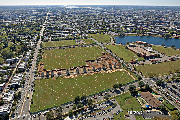 Luftaufnahme einer Stadt mit Gebäuden, Bäumen, Gras, Straßen mit Fahrzeugen, Wasser und bewölktem Himmel, die einen großen zentralen Platz zeigt, der als Parkgebiet vorgeschlagen ist.