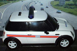 Weißes Polizeiauto mit "Polizei"-Aufschrift auf einer Straße vor einer Stadt, umgeben von fahrendem Verkehr, Gras und Bäumen auf beiden Seiten.