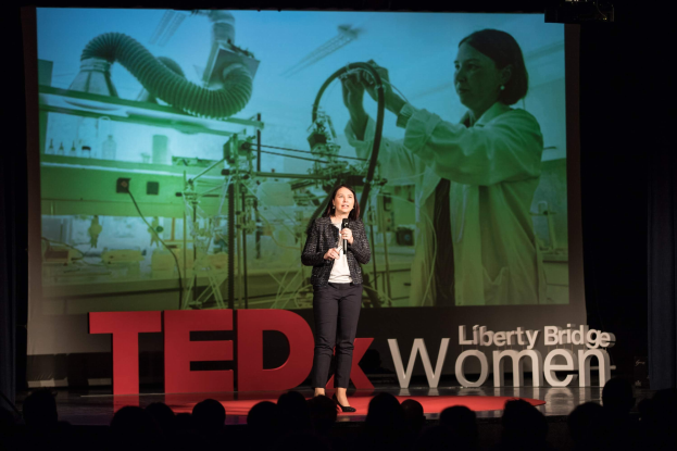 Frau auf einer Bühne mit Mikrofon, hinter ihr ein Display mit einem Bild einer Frau und Text zum TEDxWomen-Event
