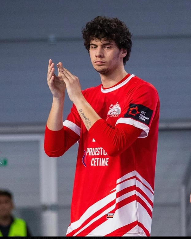 Ein Mann in einem roten und weißen Hemd klatscht in den Händen in der Vordergrund, mit einer anderen Person und einer Wand im Hintergrund und Text, der 'Kopenhagen Futsal Club - Foto 1' unten liest.