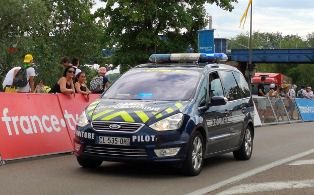 Polizeiauto fährt an einer Menschenmenge vorbei, mit einem Banner, Bäumen, einer Brücke, einer Flagge und einem bewölkten Himmel im Hintergrund.