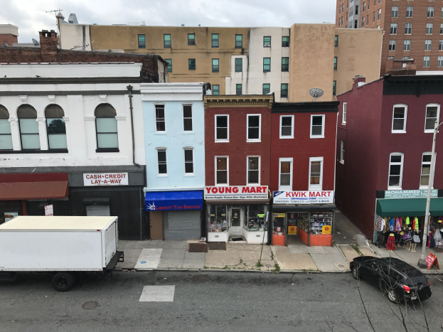 Luftaufnahme einer Stadtstraße in Brooklyn, New York, mit Fahrzeugen, Geschäftsgebäuden mit Namensschildern, Strommasten, Drähten und einem bewölkten Himmel.