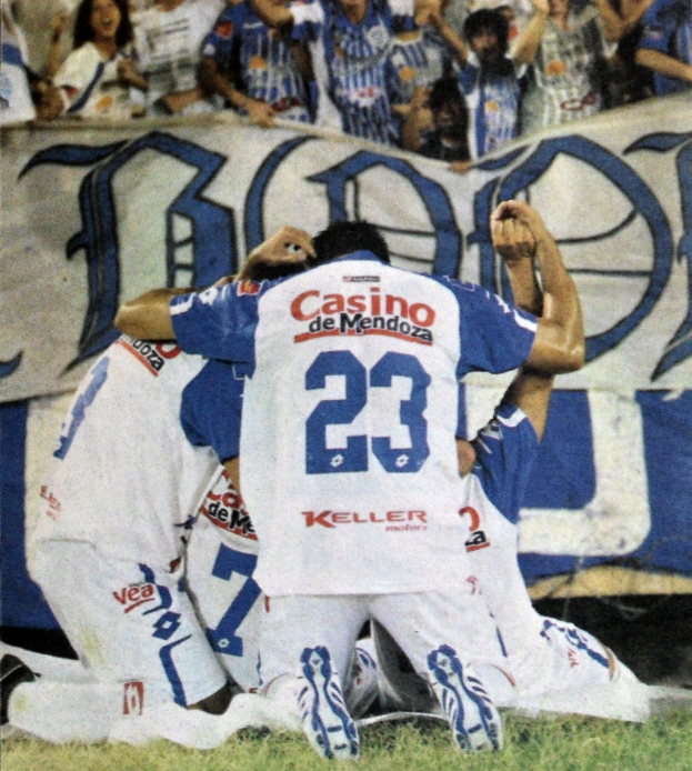 Eine Gruppe von Fußballspielern feiert ein Tor auf dem Boden vor einer jubelnden Menge, mit einem Banner mit Text im Hintergrund.