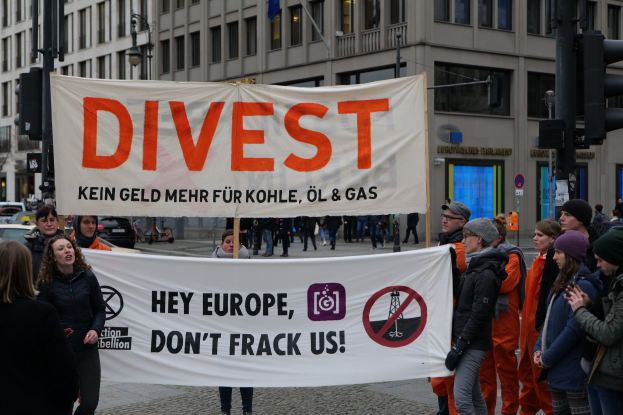 Gruppe von Menschen mit einem Banner mit der Aufschrift "Divest Hey Europe, Don't Frack Us" auf einer Straße mit Gebäuden, Straßeninfrastruktur, Fahrzeugen, Bäumen und einem klaren Himmel im Hintergrund.